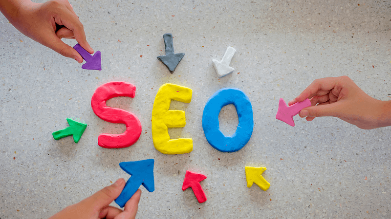 Clay letters "SEO" with colorful arrows, hands arranging them on a textured surface.